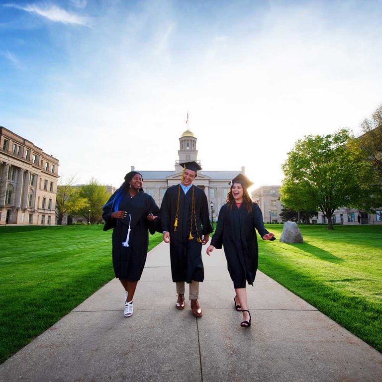 Three students in graduation cap and gowns walk in front of Old Capitol building.
