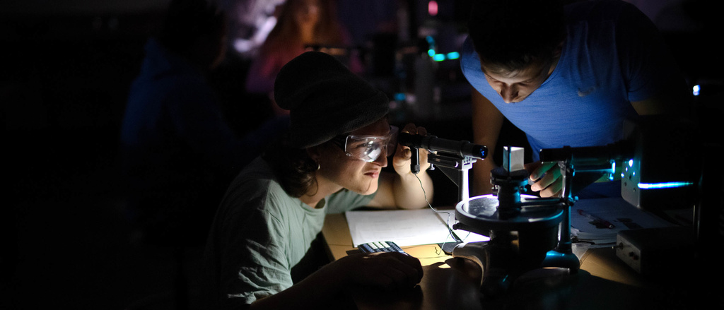 alt="A student looks through a microscope beside another student in a dark room."