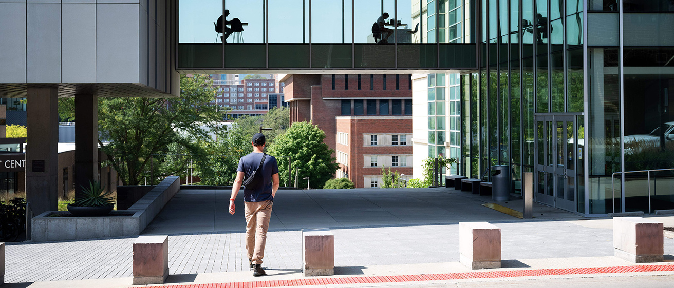 a student walking toward the engineering building, where other students are studying in the skywalk