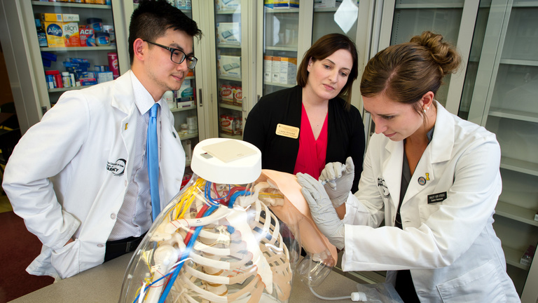 A pharmacy student practices injections on a simulated model with a professor and another student looking on.